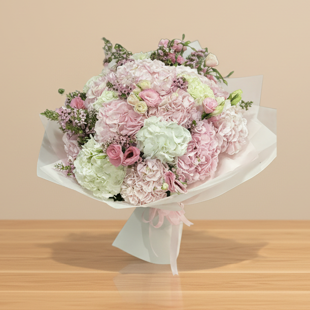 A bouquet of pink and white hydrangea flowers with various other flowers, wrapped in white paper with a pink ribbon.