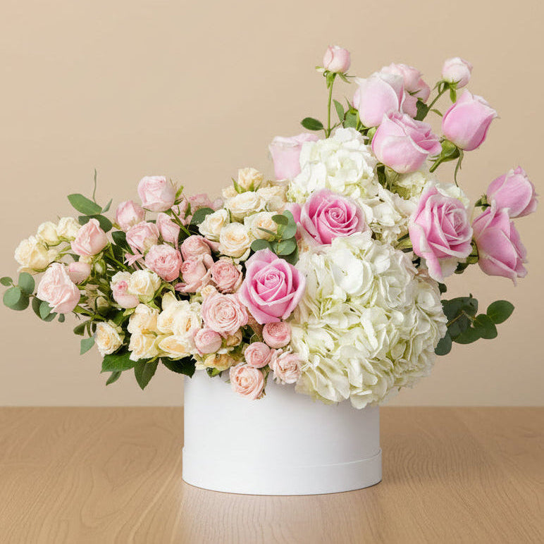 A beautiful flower arrangement in a white round box filled with white hydrangeas, pink roses, and a mix of pink and peach spray roses with fresh green eucalyptus leaves.