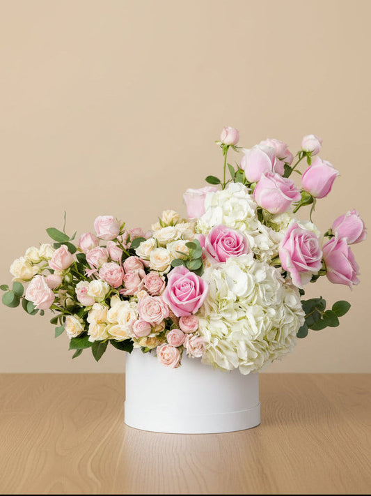 A beautiful flower arrangement in a white round box filled with white hydrangeas, pink roses, and a mix of pink and peach spray roses with fresh green eucalyptus leaves.