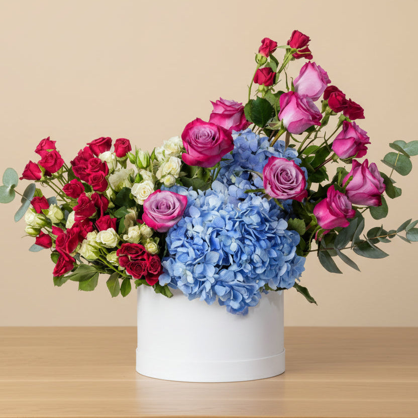 Flower arrangement in a white round box with blue hydrangea, purple roses, and red and white spray roses with green eucalyptus leaves.
