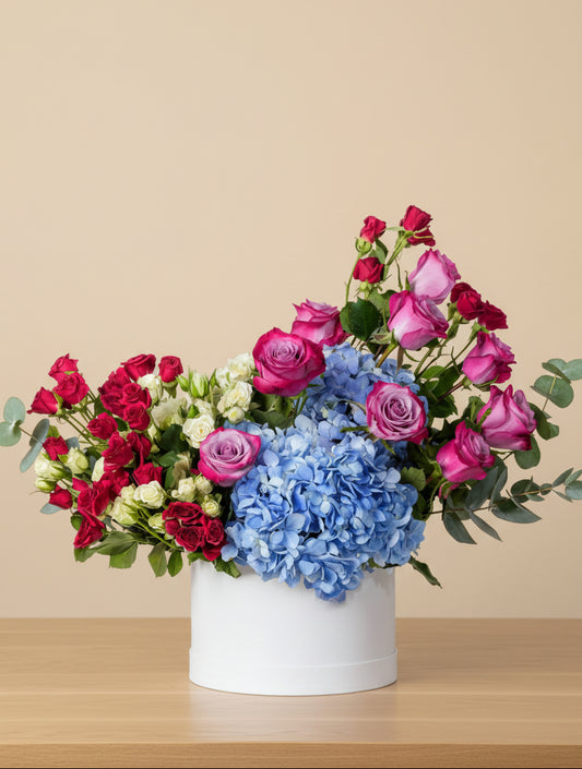 Flower arrangement in a white round box with blue hydrangea, purple roses, and red and white spray roses with green eucalyptus leaves.